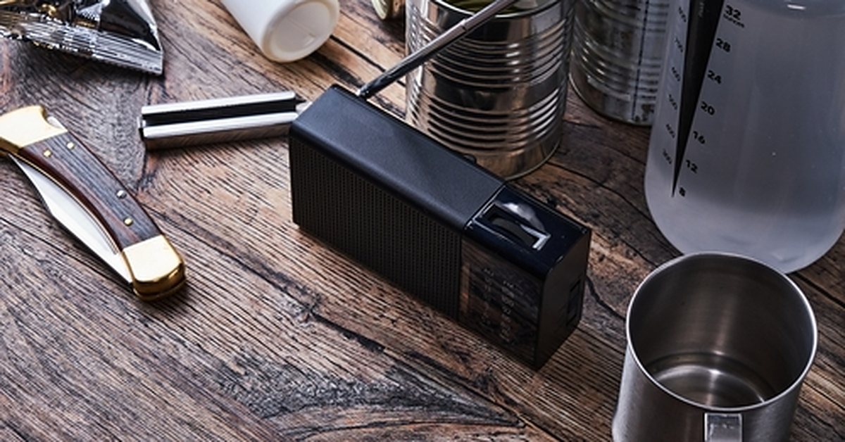 Zwarte draagbare noodradio met uitgeschoven antenne op een houten tafel, tussen conserven, een zakmes, een drinkbus en een metalen beker