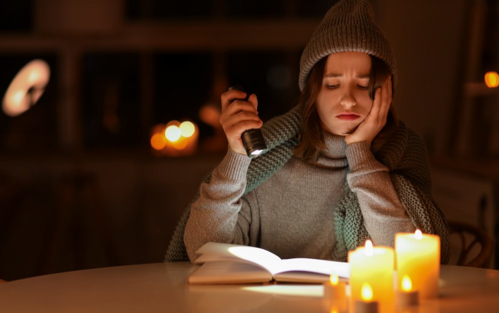 Jonge vrouw met muts en sjaal leest bij kaarslicht en een zaklamp aan tafel in een donkere keuken