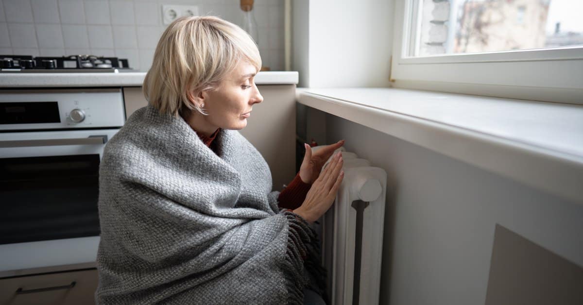 Vrouw in een deken warmt haar handen aan een radiator tijdens een verwarmingsuitval